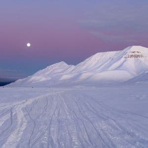Una luna piena brilla in un cielo crepuscolare violaceo sopra una vasta catena montuosa innevata con tracce nella neve.