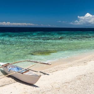Una pequeña canoa de balancín descansa en una playa de arena blanca junto a agua clara y turquesa bajo un cielo azul brillante.