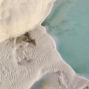 Una vista aerea di depositi minerali bianchi e testurizzati che formano terrazze accanto a uno specchio d'acqua calmo e turchese.