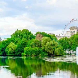 Una vista panoramica su un lago in un parco cittadino, con alberi rigogliosi e una grande ruota panoramica sullo sfondo sotto un cielo nuvoloso.