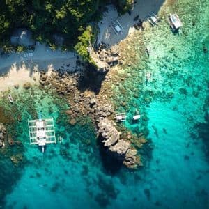 Vista aérea cenital de una cala tropical con barcos sobre agua turquesa, una playa de arena y una costa rocosa con árboles verdes.