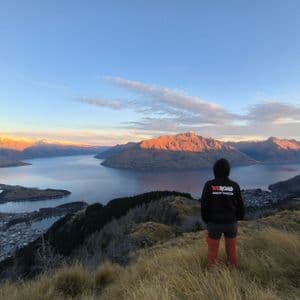 Une personne portant un sweat à capuche noir WeRoad Group Leader se tient sur une colline surplombant un vaste lac entouré de montagnes au lever du soleil.