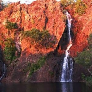 Dos cascadas caen por una pared de roca roja iluminada por el sol hacia un lago oscuro y tranquilo rodeado de árboles verdes.