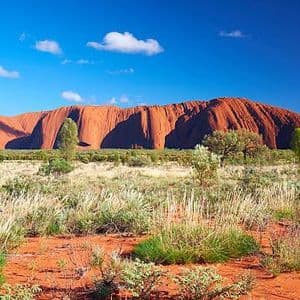 Un gran monolito de arenisca roja se eleva de una llanura de hierbas secas y arbustos bajo un cielo azul brillante con algunas nubes.