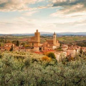 Un borgo con una torre in pietra e una chiesa è immerso tra dolci colline verdi, con ulivi in primo piano.