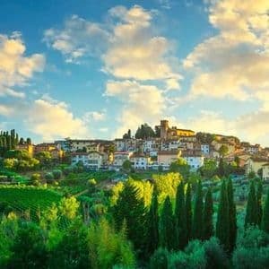 Un borgo illuminato dal sole su una collina circondata da verdi vigneti e cipressi sotto un cielo azzurro con nuvole sparse.