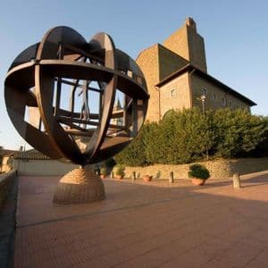 Una grande scultura sferica di metallo si erge su un piedistallo di mattoni su una terrazza pavimentata accanto a un edificio storico in pietra sotto un cielo sereno.