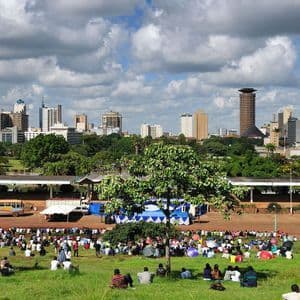 Una gran multitud de personas se sienta en el césped de un parque con un horizonte moderno de la ciudad visible al fondo bajo un cielo nublado.