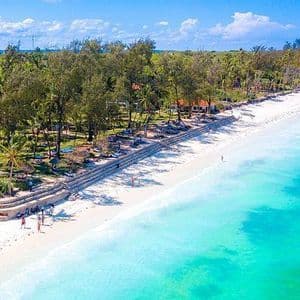 Vista aérea de una playa de arena blanca con agua turquesa, bordeada de palmeras y complejos turísticos bajo un cielo azul.