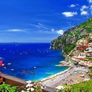 Un pueblo colorido en un acantilado verde con vistas a un mar azul salpicado de barcos, con flores brillantes en primer plano.