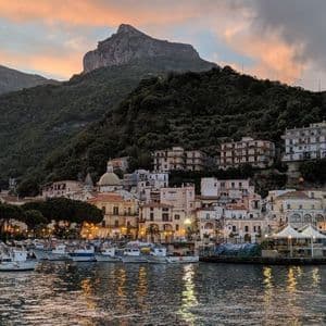 Un pueblo costero con su puerto lleno de barcos, iluminado al anochecer con una gran montaña arbolada al fondo.