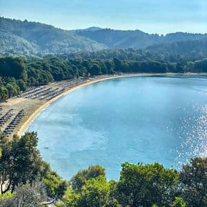 Vista dall'alto di una spiaggia sabbiosa che si estende lungo una tranquilla baia blu, con file di ombrelloni e colline boscose.
