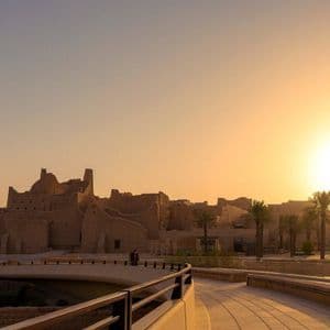 Il sole tramonta su antichi edifici di mattoni di fango, visti da una moderna passerella fiancheggiata da palme.