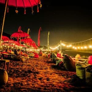 Fila di ombrelloni rossi e pouf su una spiaggia sabbiosa di notte, illuminati da calde lucine.
