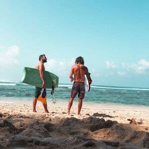 Due uomini di un viaggio di gruppo WeRoad si trovano su una spiaggia sabbiosa con bodyboard, guardando l'oceano sotto un cielo azzurro.