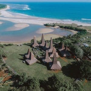 Una vista aerea di un villaggio tradizionale con alti tetti di paglia su una collina erbosa che si affaccia su una spiaggia, una laguna e l'oceano.