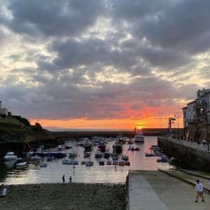 Un porto pieno di piccole barche al tramonto, con un sole arancione basso all'orizzonte sotto un cielo molto nuvoloso.
