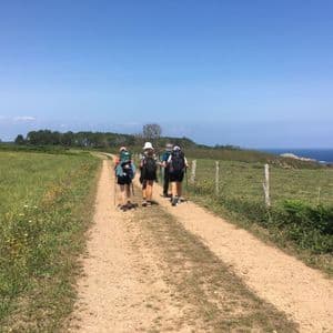 Un viaggio di gruppo WeRoad in escursione con zaini e bastoncini su un sentiero sterrato lungo una costa erbosa sotto un cielo azzurro e limpido.