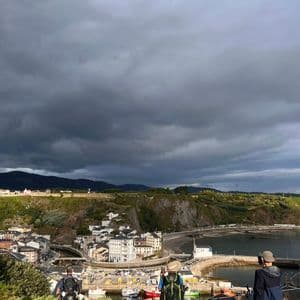 Un gruppo WeRoad di tre escursionisti con zaini che domina una cittadina portuale costiera da un punto panoramico sotto un cielo nuvoloso e drammatico.