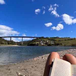 Una persona seduta su una riva sabbiosa legge un libro, con un fiume, colline verdi e un ponte sullo sfondo.