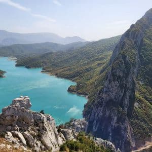 Una vista dall'alto da un crinale montuoso roccioso che si affaccia su un sinuoso lago turchese circondato da verdi colline boscose.