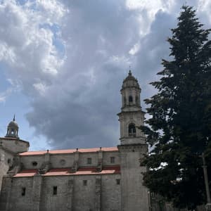 Una chiesa in pietra con campanile e tetto in tegole rosse si erge accanto a un alto albero sempreverde sotto un cielo drammatico e nuvoloso.