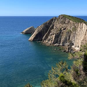 Una vista dall'alto di un promontorio roccioso che si estende in un mare calmo e turchese con una piccola caletta sottostante.