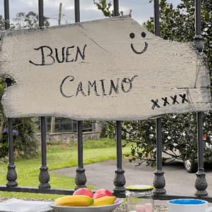 Un cartello di legno con la scritta 'Buen Camino' è appeso a una recinzione, sopra un muro di pietra che contiene una ciotola di frutta e un barattolo di dolci.