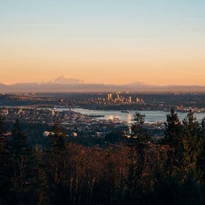 Vue plongeante sur une silhouette urbaine au bord de l'eau au lever du soleil, avec des montagnes enneigées au loin et la cime des arbres en contrebas.