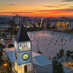 Una vista aerea di un'ampia piazza cittadina con una torre dell'orologio in primo piano e persone che passeggiano durante un tramonto colorato.