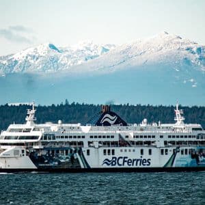 Un grand navire blanc de BC Ferries navigue sur l'eau, avec un rivage boisé et des montagnes enneigées en arrière-plan.