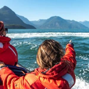 Deux personnes d'un voyage en groupe WeRoad, vêtues de vestes rouges, sur un bateau, l'une d'elles pointant du doigt des montagnes lointaines.