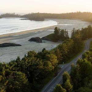 Vue aérienne d'une route sinueuse traversant une forêt luxuriante et verdoyante le long d'une large plage de sable au coucher du soleil.