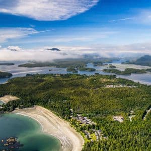 Vue aérienne d'un archipel boisé avec une plage de sable incurvée, entouré d'eau et de petites îles sous un ciel partiellement nuageux.
