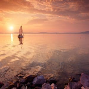 Ein Segelboot gleitet auf ruhigem Wasser während eines leuchtenden orangefarbenen Sonnenuntergangs, dessen Spiegelung auf der Oberfläche sichtbar ist, mit Bergen in der Ferne.