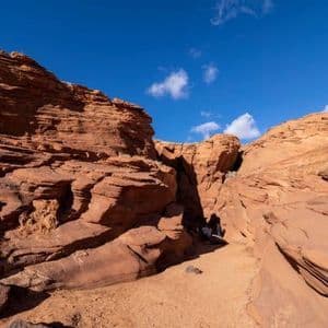 Dos personas de un viaje en grupo de WeRoad descansan a la sombra de un estrecho sendero de cañón de arenisca bajo un cielo azul brillante.
