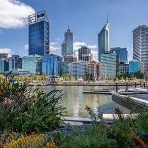 Un parque frente al mar con arbustos verdes y flores que da a un moderno horizonte urbano con rascacielos bajo un cielo parcialmente nublado.