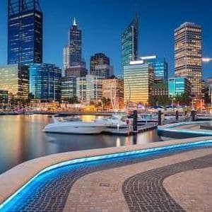 Un horizonte urbano moderno con rascacielos iluminados al anochecer, reflejándose en un puerto deportivo con barcos amarrados y un paseo marítimo curvo e iluminado.