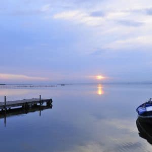 Un barco azul está amarrado a un muelle de madera en un lago en calma al atardecer, con el reflejo del sol en el agua.