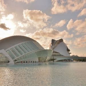 Dos grandes edificios blancos modernistas con fachadas de cristal situados junto a una gran lámina de agua bajo un cielo nublado al atardecer.