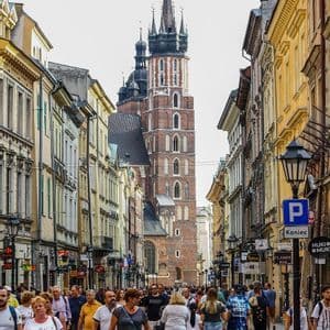 Una multitud de personas camina por una estrecha calle histórica, flanqueada por edificios antiguos, con una gran torre de iglesia de ladrillo al fondo.