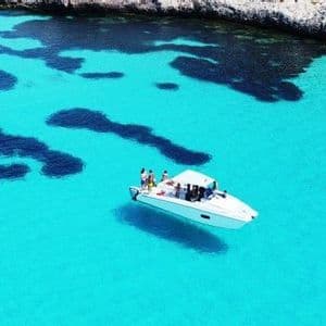Una vista aerea di un viaggio di gruppo WeRoad su un motoscafo bianco che galleggia in un mare calmo e turchese vicino a una costa rocciosa.