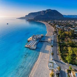 Vista aerea di una lunga spiaggia sabbiosa con un molo ornato che si protende nel mare turchese, accanto a una città ai piedi di una montagna.