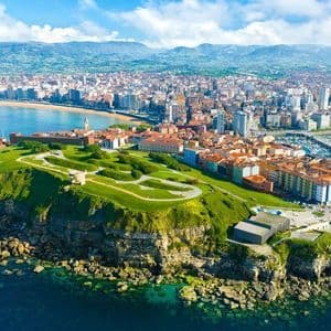 Una vista aérea de una ciudad costera con una península verde, un gran puerto deportivo con barcos y una playa de arena.