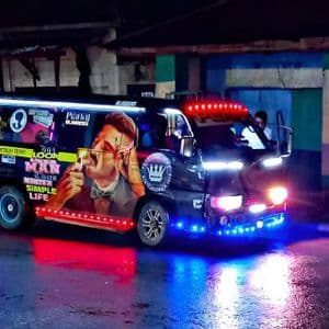 Un minibús brillantemente decorado con luces LED azules y rojas se encuentra en una calle mojada de la ciudad por la noche, sus luces reflejándose en el pavimento.