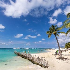 Une plage tropicale ensoleillée avec des palmiers, du sable blanc et une eau turquoise. Des gens se détendent et nagent, avec des bateaux ancrés au large.