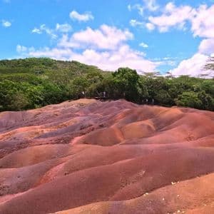 Des dunes ondulantes de terre multicolore aux nuances de rouge et de brun se détachent sur un fond de forêt verdoyante sous un ciel bleu parsemé de nuages.