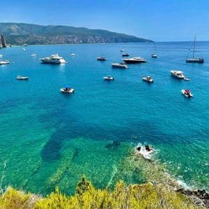 Veduta dall'alto di numerose imbarcazioni e yacht su acque limpide e turchesi in una baia costiera con scogliere sullo sfondo.