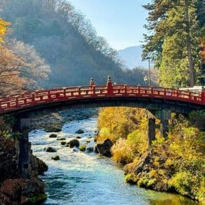 Un tradizionale ponte ad arco rosso attraversa un fiume limpido e roccioso in una valle con alberi autunnali colorati.