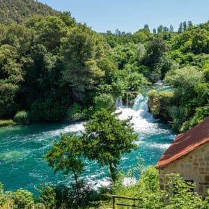 Une maison en pierre au toit de tuiles rouges surplombe une rivière turquoise et des cascades s'écoulant à travers une forêt verdoyante et luxuriante.